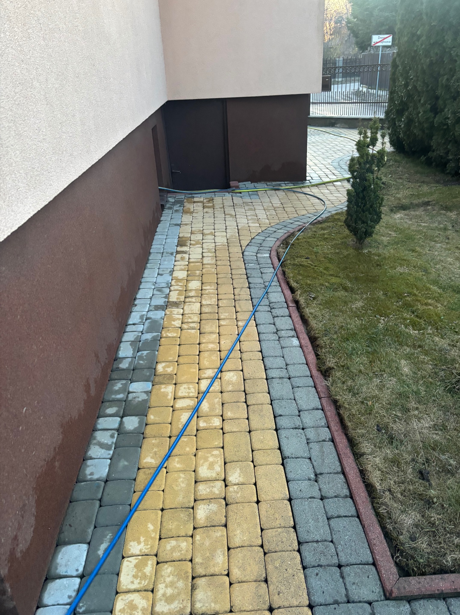 Częściowo umyta, dwukolorowa kostka brukowa (żółta i szara) przed budynkiem. Widoczny wąż ciśnieniowy i fragment trawnika z tujami. Efekt czyszczenia.