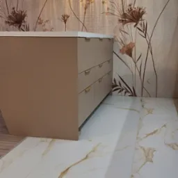 Beżowa komoda z złotymi uchwytami na tle ściany w kwiaty i marmurowej podłogi. Dwie wiszące lampy kuliste oświetlają wnętrze.