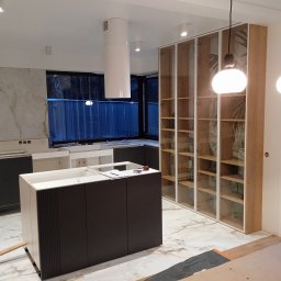 Wnętrze w trakcie montażu: wyspa kuchenna z ciemnymi frontami i białą ramą, przeszklona szafa z drewnianymi półkami, marmurowa podłoga, okap cylindryczny.