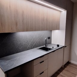 Nowoczesna kuchnia z drewnianymi szafkami górnymi podświetlonymi LED, czarnym blatem i zlewozmywakiem. Minimalistyczne uchwyty i drewniana podłoga.