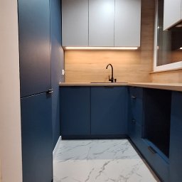 Niebiesko-szara kuchnia z drewnianym blatem i marmurową podłogą. Minimalistyczny design, czarna bateria, oświetlenie LED pod szafkami. Nowoczesne wykończenie.