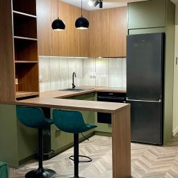 Nowoczesna kuchnia z drewnianymi frontami, zielonymi akcentami i wyspą z hokerami. Uwagę przykuwa czarna lodówka i designerskie lampy wiszące.