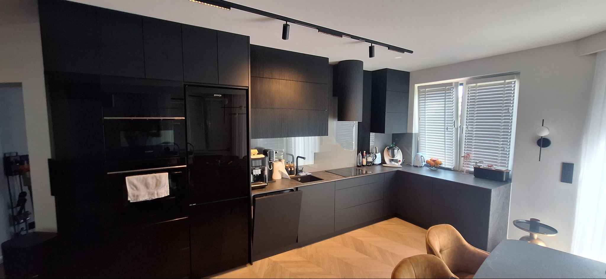 Nowoczesna, czarna kuchnia na wymiar z frezowanymi frontami, oświetleniem punktowym i drewnianą podłogą w jodełkę. Widok na okno z żaluzjami.