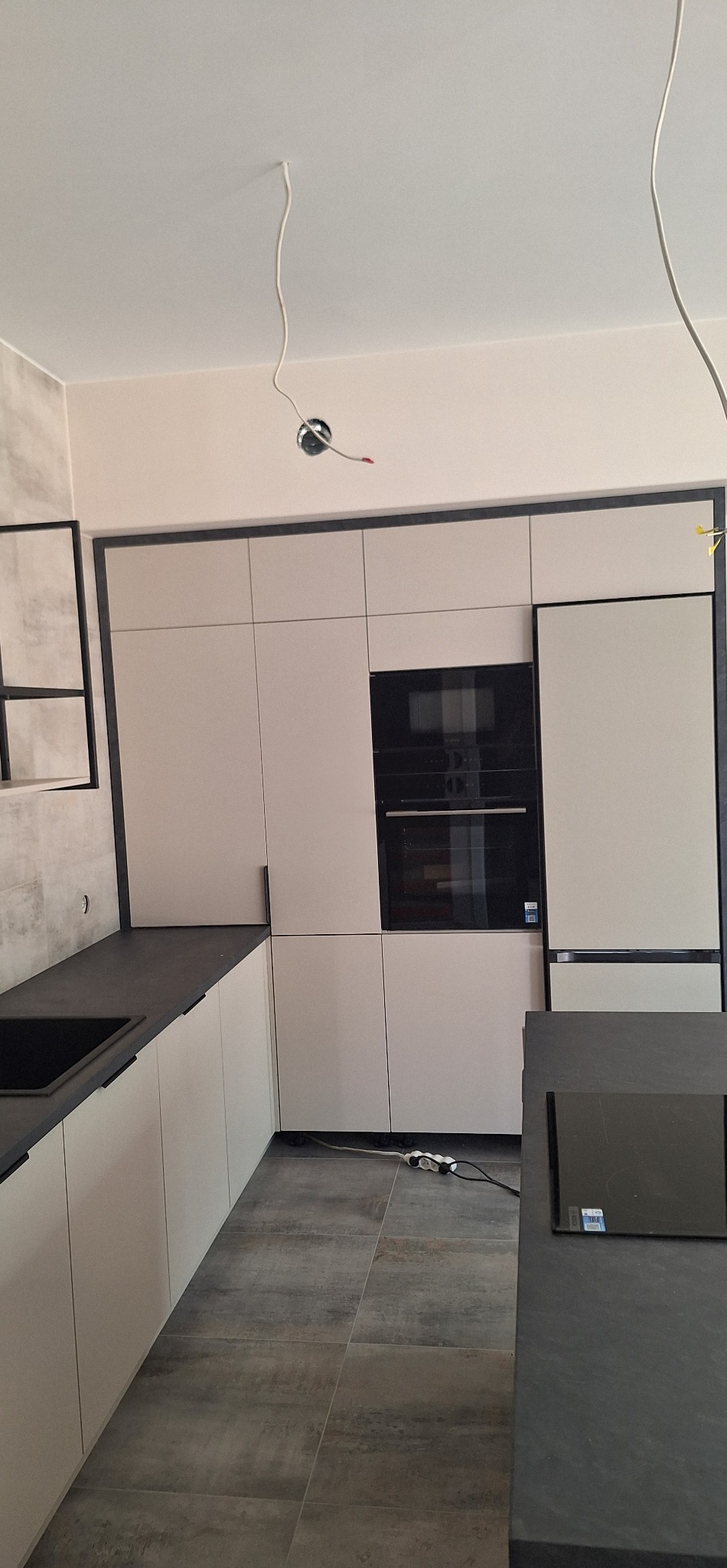 Jasna zabudowa kuchenna z czarnymi akcentami: blat, obramowania. Widoczne AGD w zabudowie. Na podłodze szare płytki imitujące beton. W tle wystające kable.