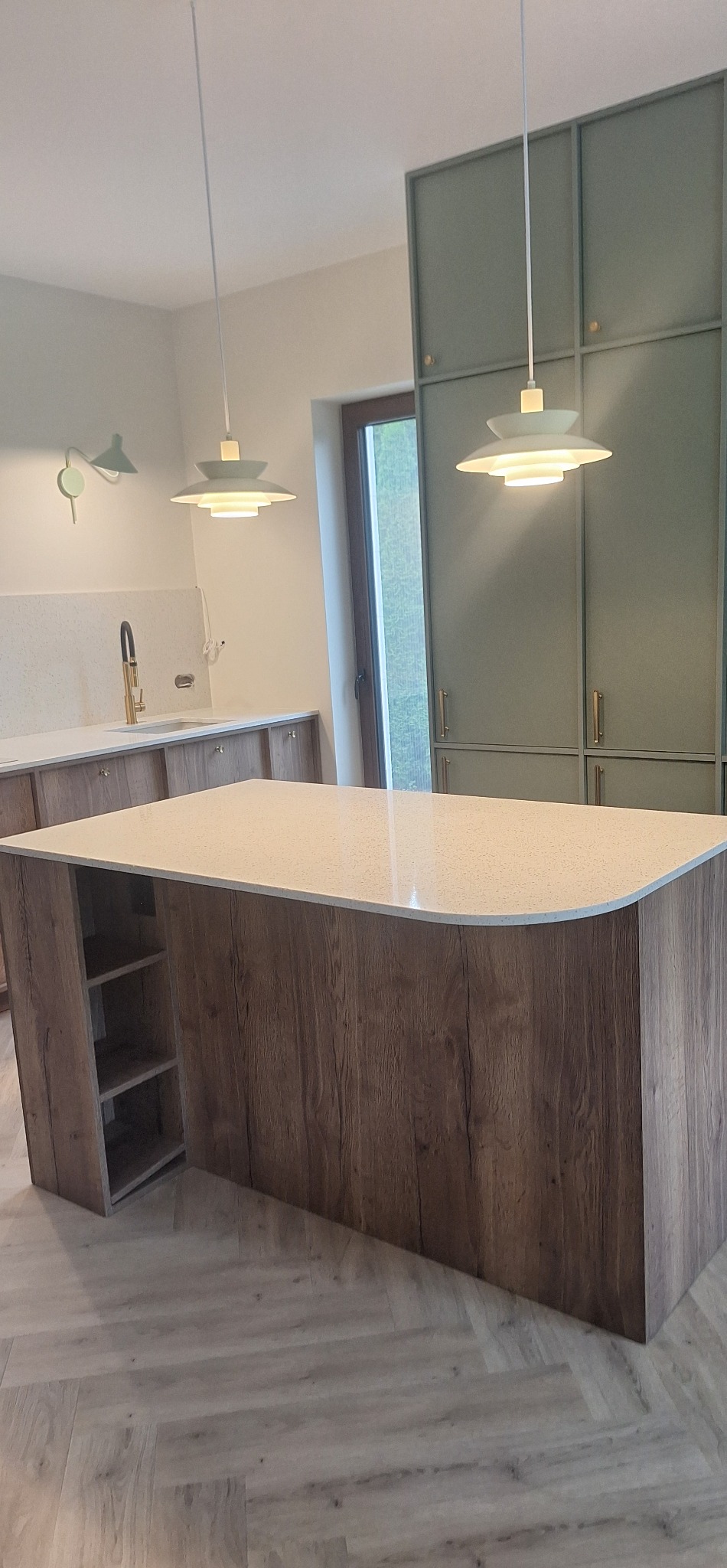 Nowoczesna wyspa kuchenna z drewnianym wykończeniem i jasnym blatem, oświetlona wiszącymi lampami. W tle zabudowa kuchenna w odcieniach zieleni i parkiet w jodełkę.