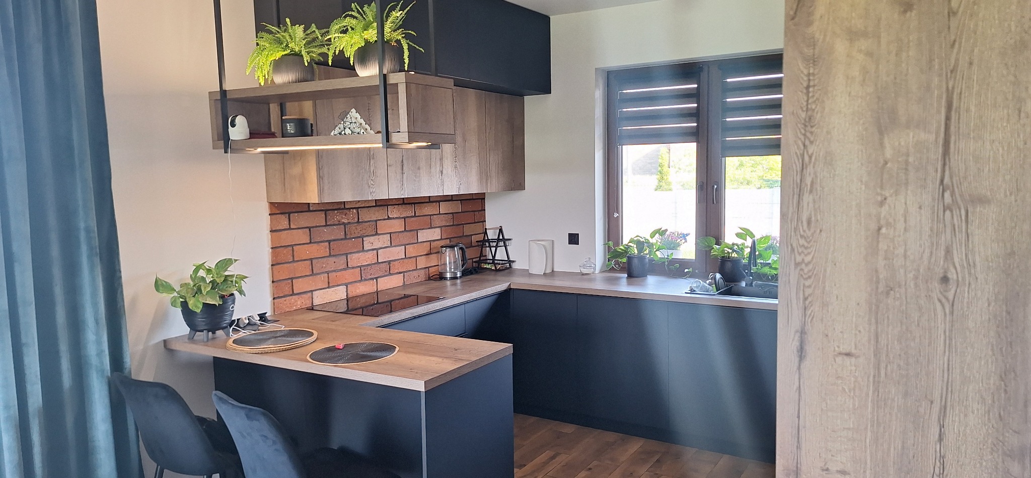 Nowoczesna kuchnia z czarnymi szafkami i drewnianymi blatami. Na ścianie cegła, okno z roletami, rośliny doniczkowe, oświetlenie podszafkowe.