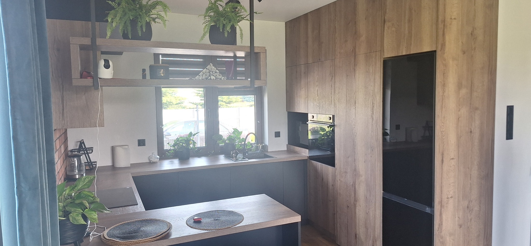Nowoczesna kuchnia z drewnianymi frontami i czarnymi blatami, oświetlona naturalnym światłem z okna. Rośliny doniczkowe dodają wnętrzu świeżości i charakteru.