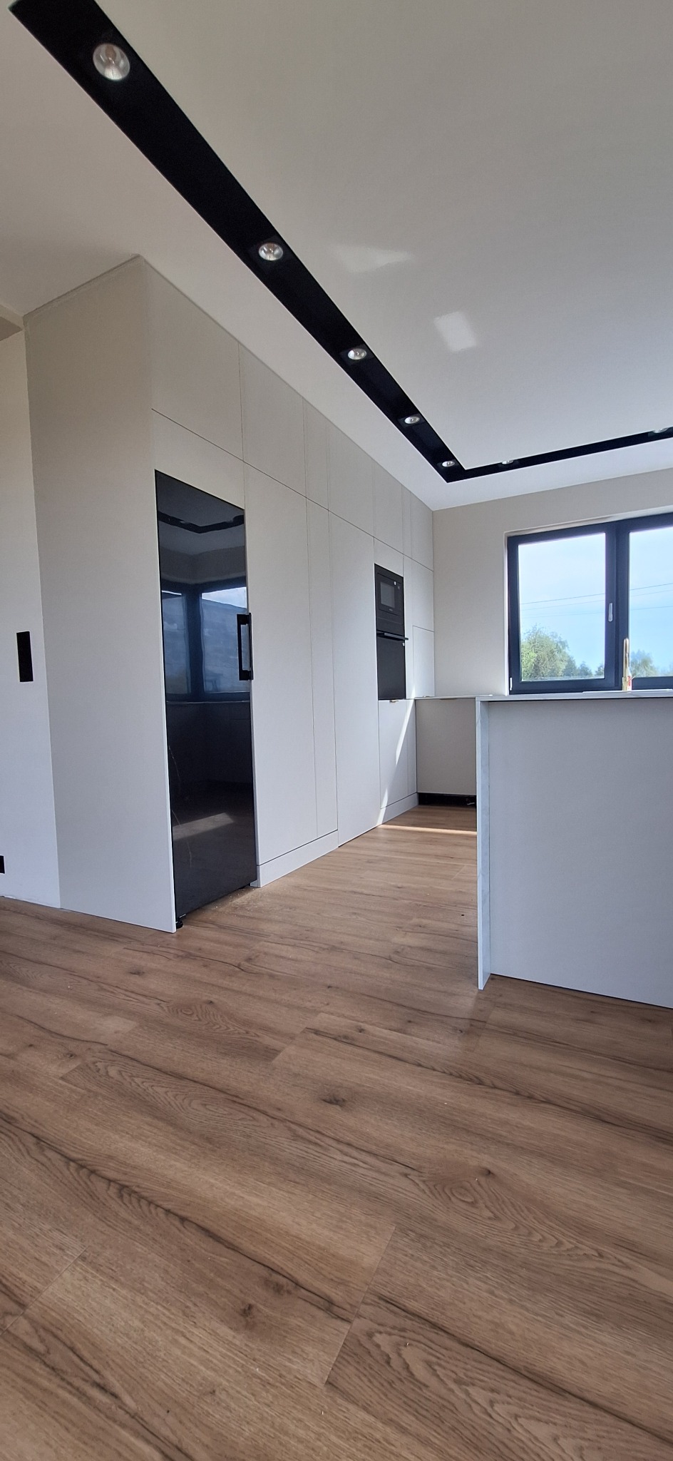 Nowoczesna kuchnia z białymi szafkami, czarnym sprzętem AGD i drewnianą podłogą. W tle okno z widokiem na zieleń. Oświetlenie punktowe.