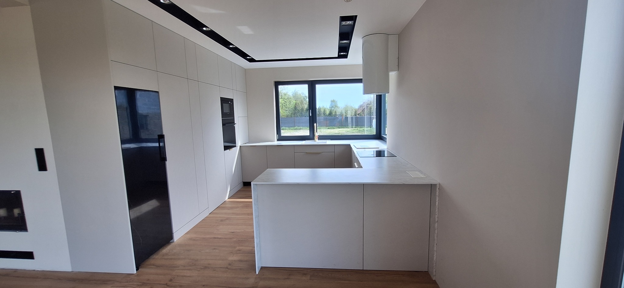 Jasna kuchnia z wyspą i czarnymi akcentami. Nowoczesne meble kuchenne z zabudowaną lodówką i piekarnikiem w minimalistycznym stylu, z widokiem na zieleń za oknem.