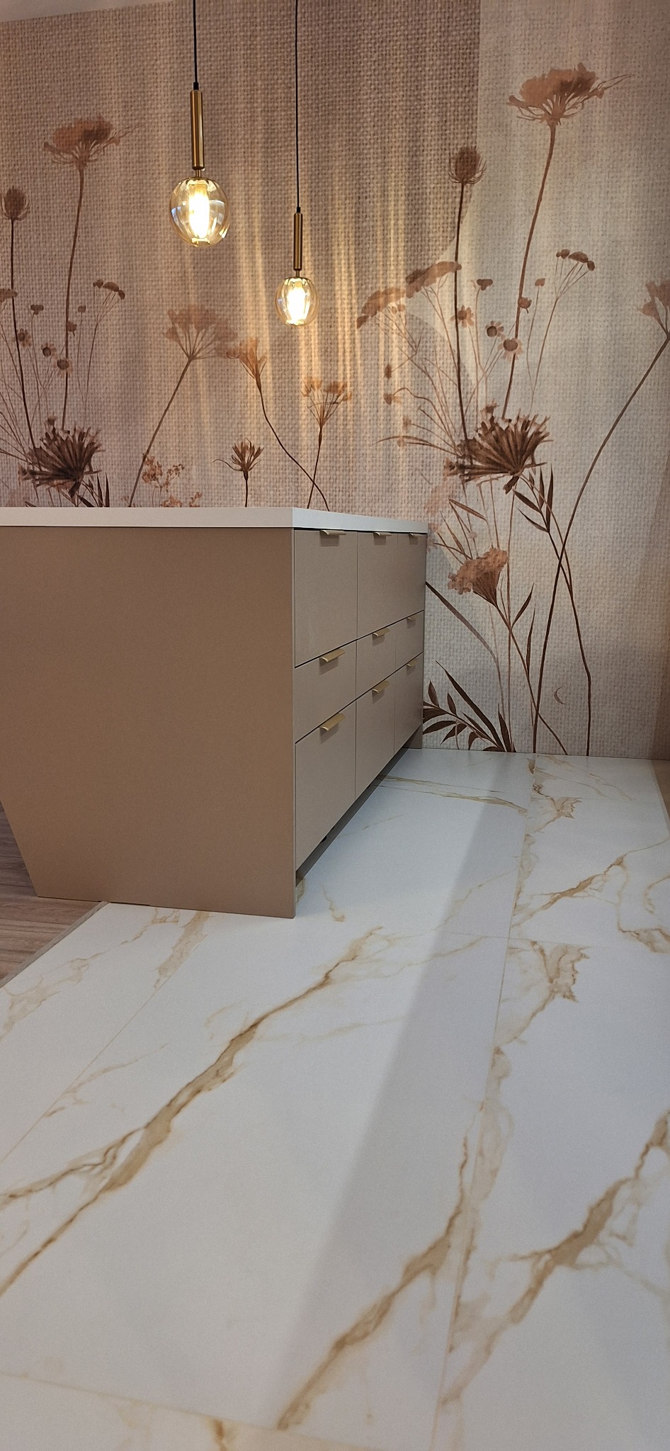 Beżowa komoda z złotymi uchwytami na tle ściany w kwiaty i marmurowej podłogi. Dwie wiszące lampy kuliste oświetlają wnętrze.