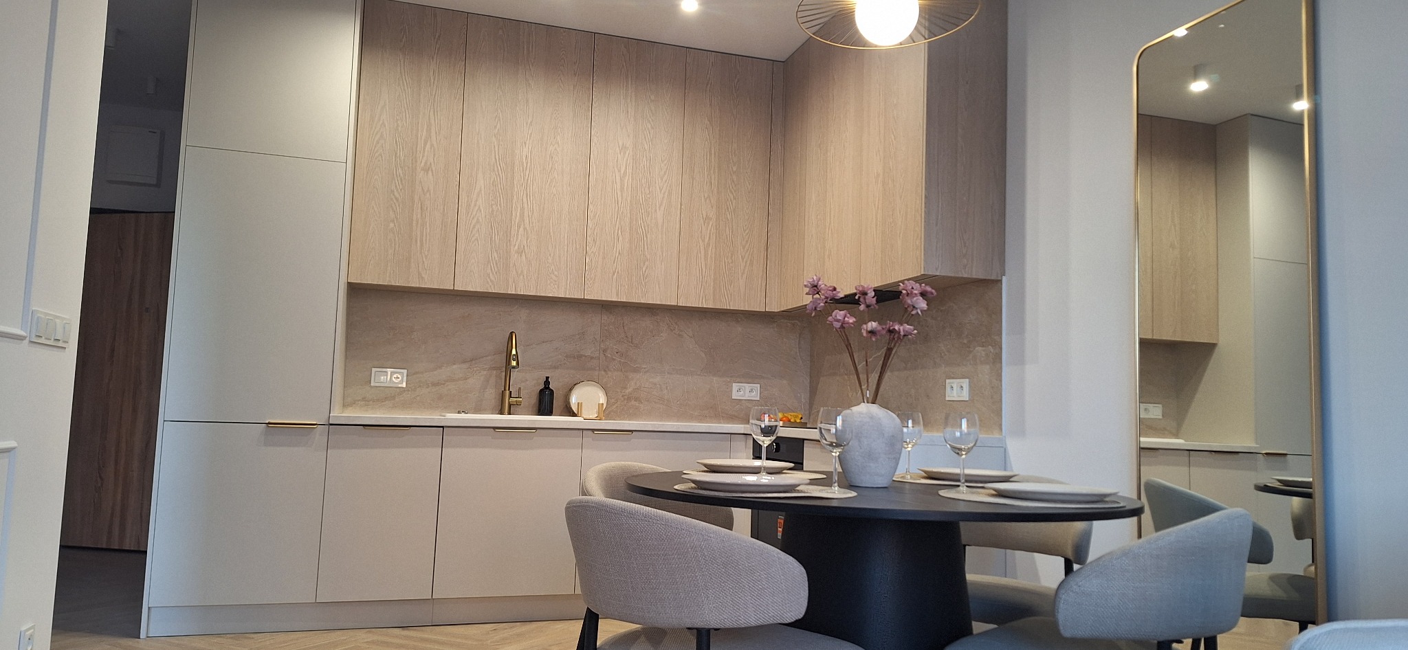 Jasna kuchnia z drewnianymi frontami szafek, złotą armaturą i okrągłym stołem jadalnym. W tle lustro odbijające fragment kuchni.