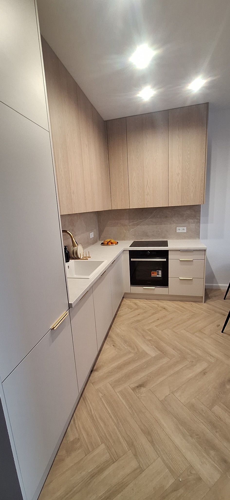 Jasna kuchnia w stylu skandynawskim z drewnianymi frontami szafek i złotymi uchwytami, wykończona parkietem w jodełkę. Widok na płytę indukcyjną i owoce na blacie.