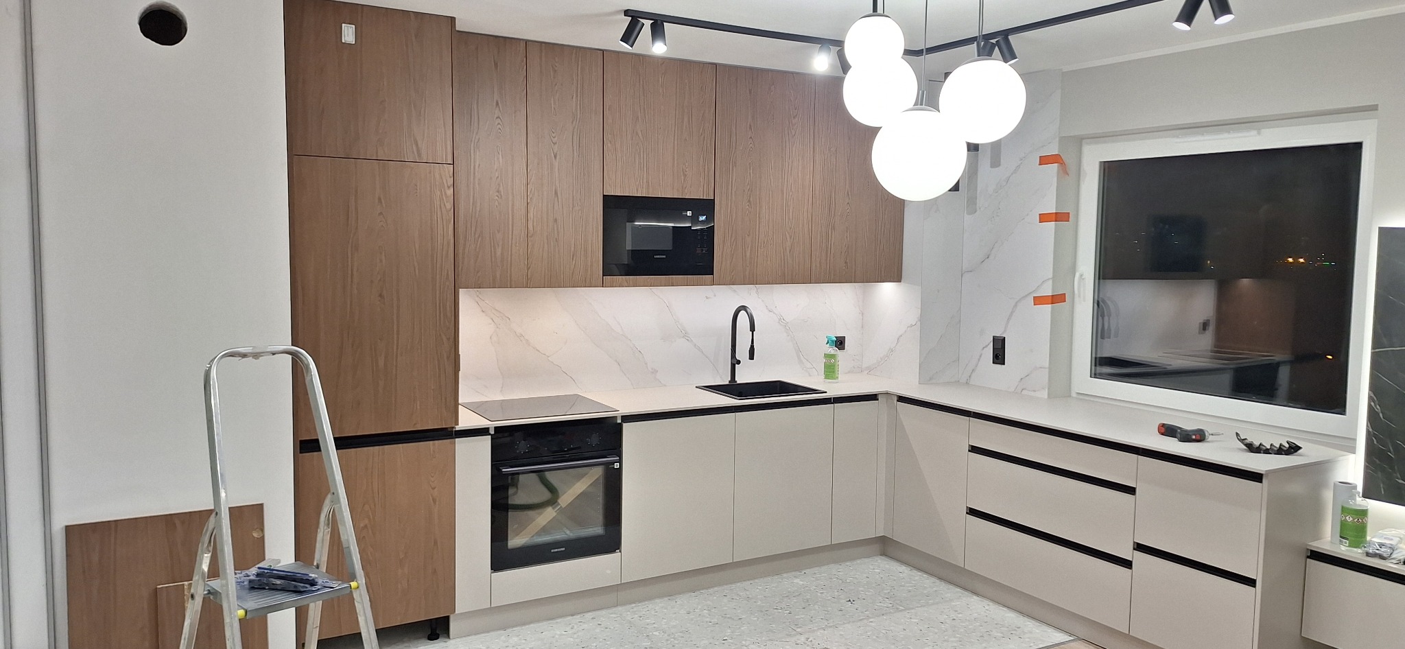 Nowoczesna kuchnia z drewnianymi szafkami górnymi i beżowymi dolnymi. Na blacie czarny zlew i płyta. Widoczna drabina malarska. Styl minimalistyczny, oświetlenie punktowe.