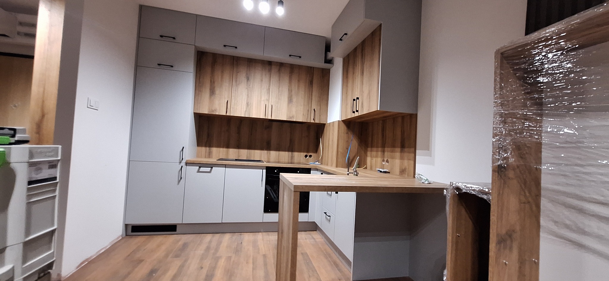 Nowoczesna kuchnia z szarymi frontami i drewnianymi akcentami, oświetlenie punktowe, wyspa kuchenna z blatem w kolorze drewna, fragment ramy w folii ochronnej.
