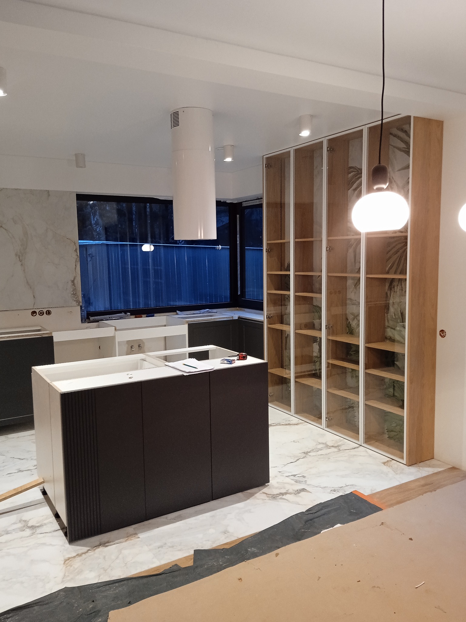 Wnętrze w trakcie montażu: wyspa kuchenna z ciemnymi frontami i białą ramą, przeszklona szafa z drewnianymi półkami, marmurowa podłoga, okap cylindryczny.
