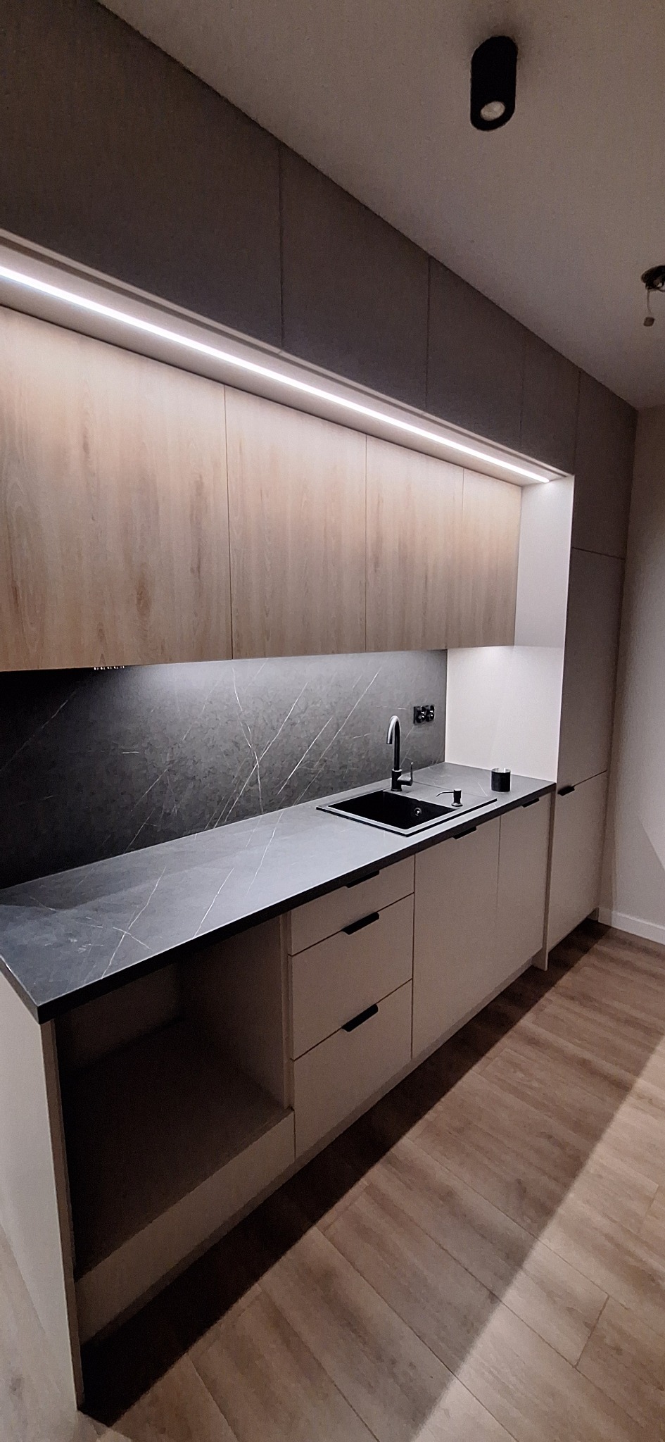 Nowoczesna kuchnia z drewnianymi szafkami górnymi podświetlonymi LED, czarnym blatem i zlewozmywakiem. Minimalistyczne uchwyty i drewniana podłoga.