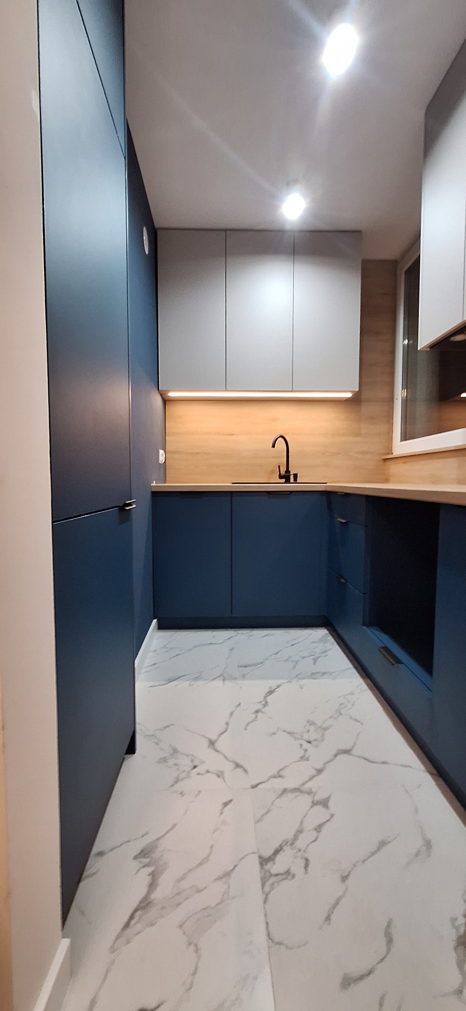 Niebiesko-szara kuchnia z drewnianym blatem i marmurową podłogą. Minimalistyczny design, czarna bateria, oświetlenie LED pod szafkami. Nowoczesne wykończenie.