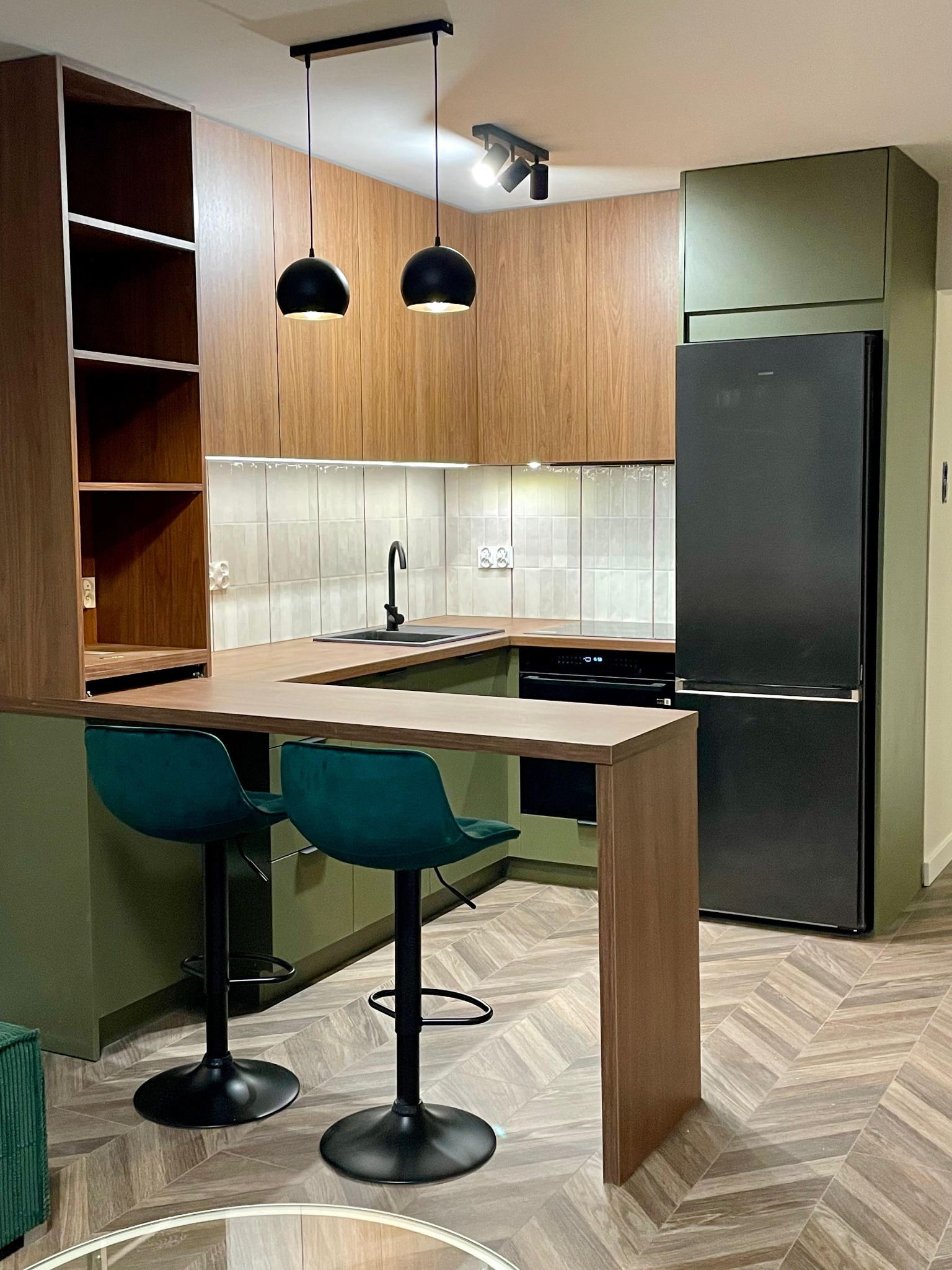 Nowoczesna kuchnia z drewnianymi frontami, zielonymi akcentami i wyspą z hokerami. Uwagę przykuwa czarna lodówka i designerskie lampy wiszące.