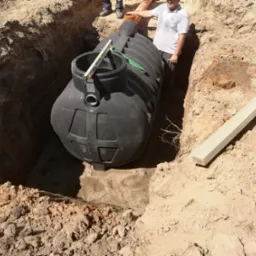 Czarny zbiornik na szambo umieszczony w wykopie w Toruniu, poziomica na górze zbiornika, widoczny fragment pracownika firmy instalacyjnej.