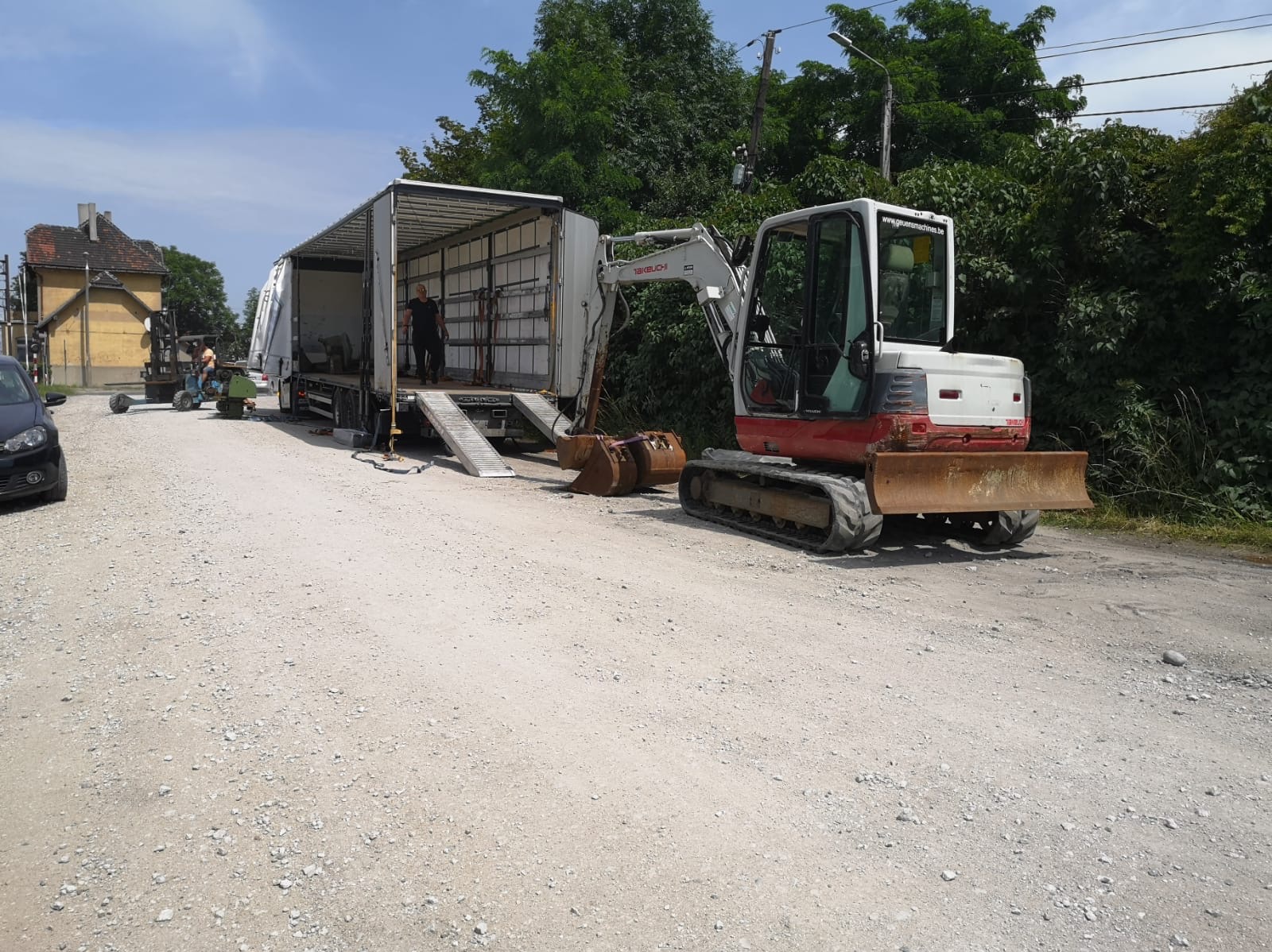 Mała koparka gąsienicowa i ciężarówka z rampą na żwirowej drodze, widoczny operator i budynek w tle.