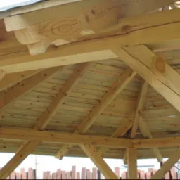 Drewniana konstrukcja dachu altany z widocznymi elementami dekoracyjnymi, wykonana z jasnego drewna, z widocznym deskowaniem i fragmentem pokrycia dachowego.