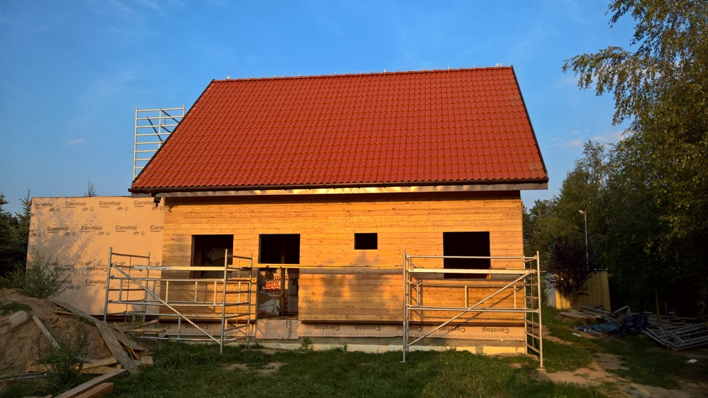 Surowy stan domu szkieletowego z czerwoną dachówką, widoczne rusztowania i folia izolacyjna na ścianach zewnętrznych.