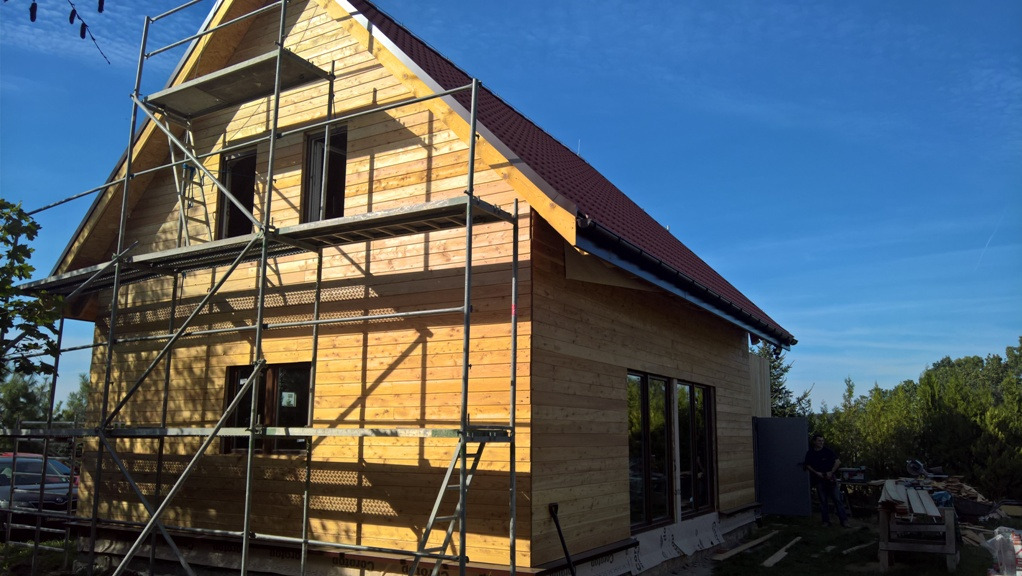 Surowy dom szkieletowy z drewna, częściowo pokryty czerwoną dachówką, otoczony rusztowaniami i przygotowany do dalszych prac wykończeniowych, widoczny fragment ogrodu i błękitne niebo.