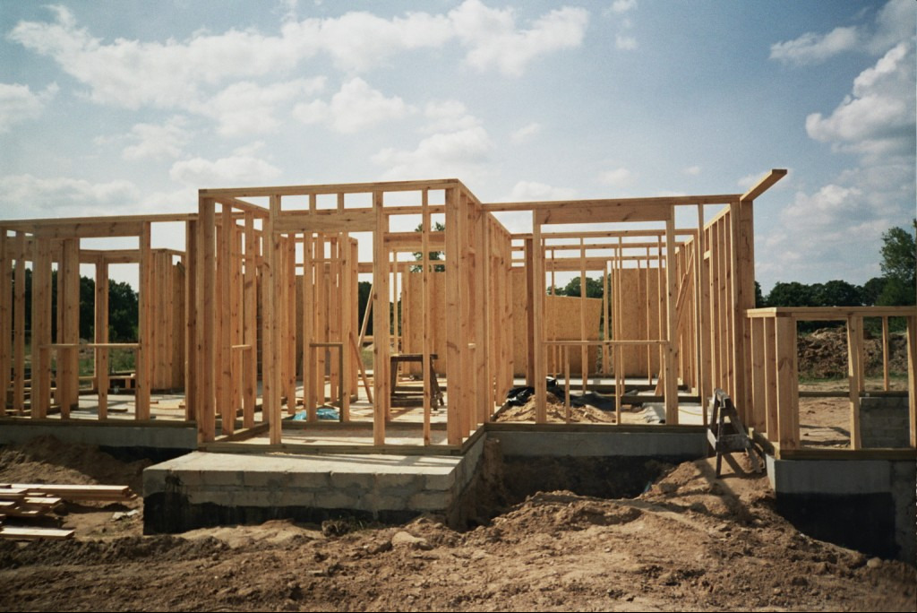 Surowa konstrukcja domu szkieletowego z jasnego drewna na betonowym fundamencie, widok z zewnątrz, w tle drzewa i pochmurne niebo.