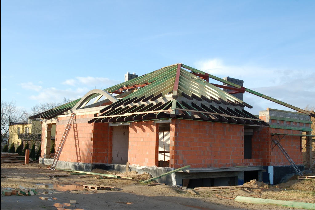 Niedokończony dom z czerwonej cegły z widoczną drewnianą konstrukcją dachu i łukowym oknem, drabiny opierają się o ściany, palety leżą na ziemi.