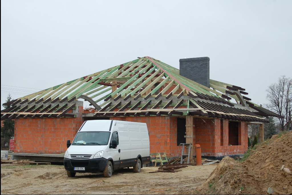 Budowa domu jednorodzinnego w trakcie wznoszenia więźby dachowej, widoczny biały samochód dostawczy Iveco na placu budowy.