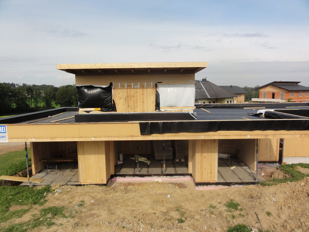 Wznoszenie domu szkieletowego z drewnianą konstrukcją, widoczna izolacja w workach i czarna membrana dachowa, w tle zabudowa jednorodzinna.