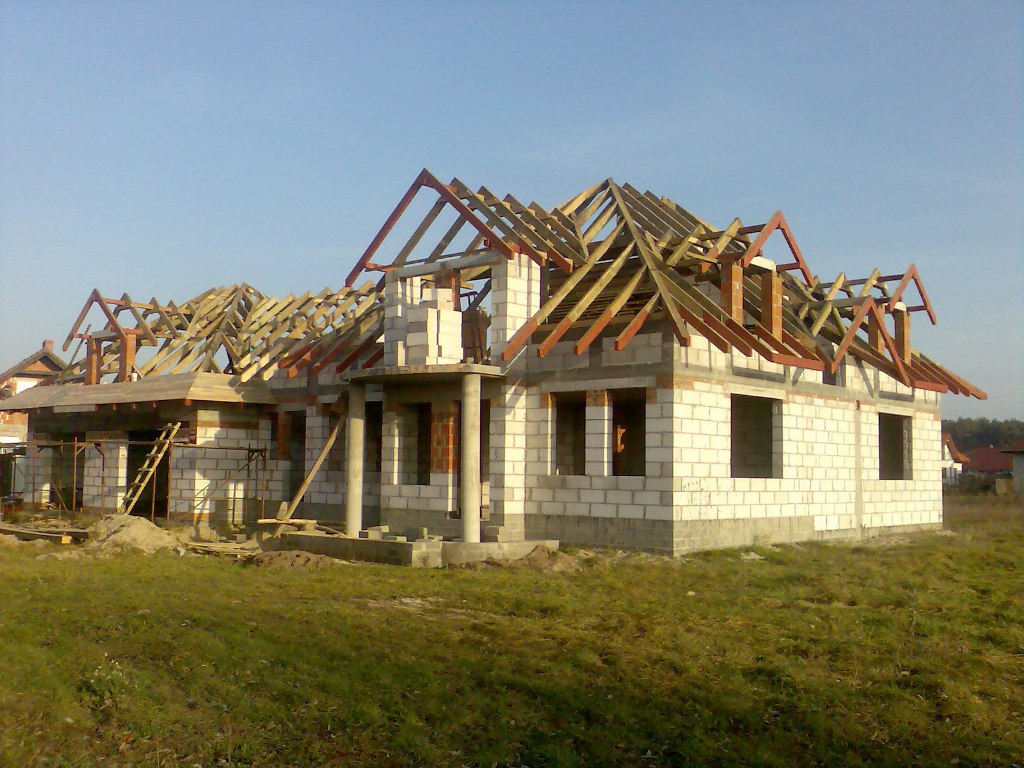 Budowa domu jednorodzinnego w stanie surowym otwartym, widoczna konstrukcja ścian z bloczków oraz drewniana konstrukcja dachu, w tle błękitne niebo.