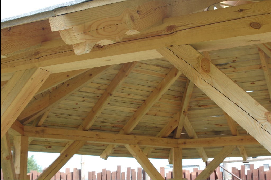 Drewniana konstrukcja dachu altany z widocznymi elementami dekoracyjnymi, wykonana z jasnego drewna, z widocznym deskowaniem i fragmentem pokrycia dachowego.