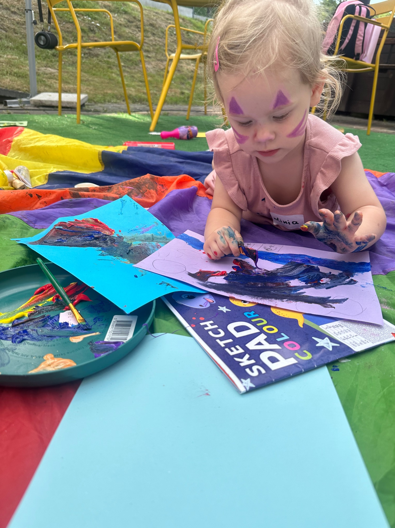 Dziewczynka z pomalowaną twarzą i imieniem 'Vinia' na koszulce, leżąca na kolorowej płachcie i malująca farbami na kartkach papieru, obok paleta z farbami i blok rysunkowy.