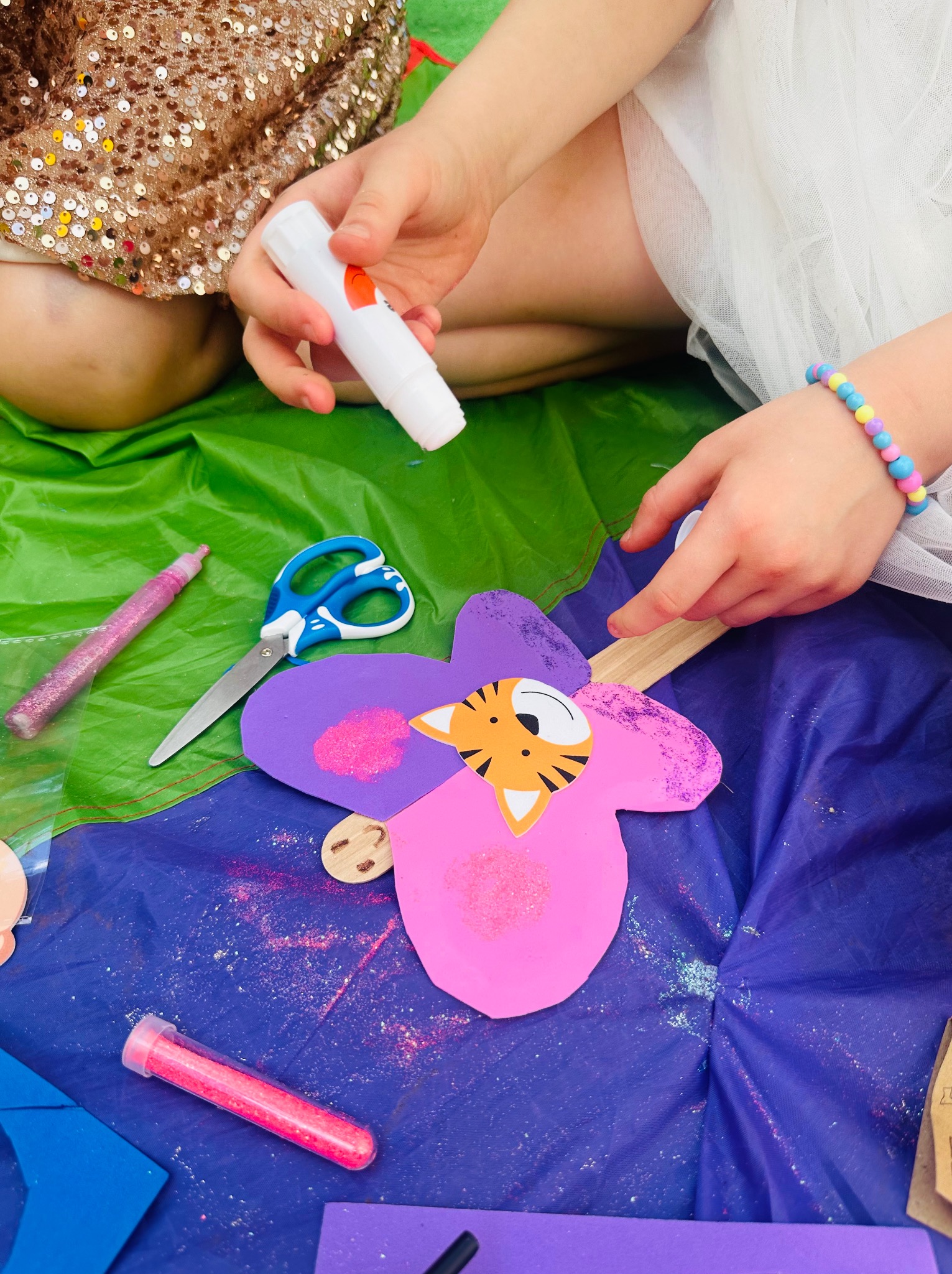 Dziecko w sukience z cekinami i dziecko w białej tiulowej sukience tworzą kolorowego kwiatka z papieru z motywem tygrysa, używając kleju w sztyfcie i brokatu na zielonym i niebieskim tle.