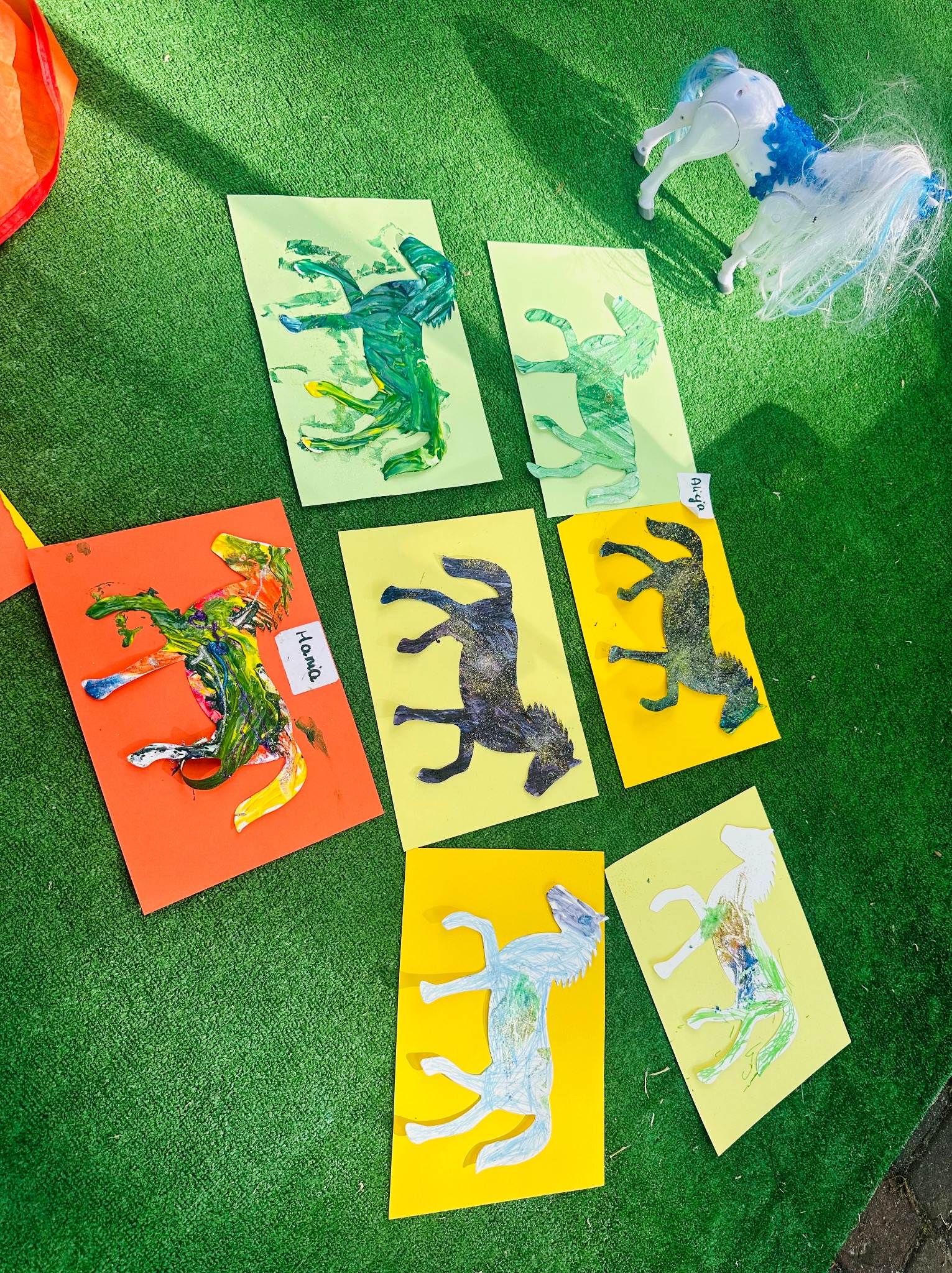 Ręcznie malowane farbami konie na kolorowych kartkach, leżące na zielonej sztucznej trawie, obok zabawkowy plastikowy koń z niebieską grzywą.