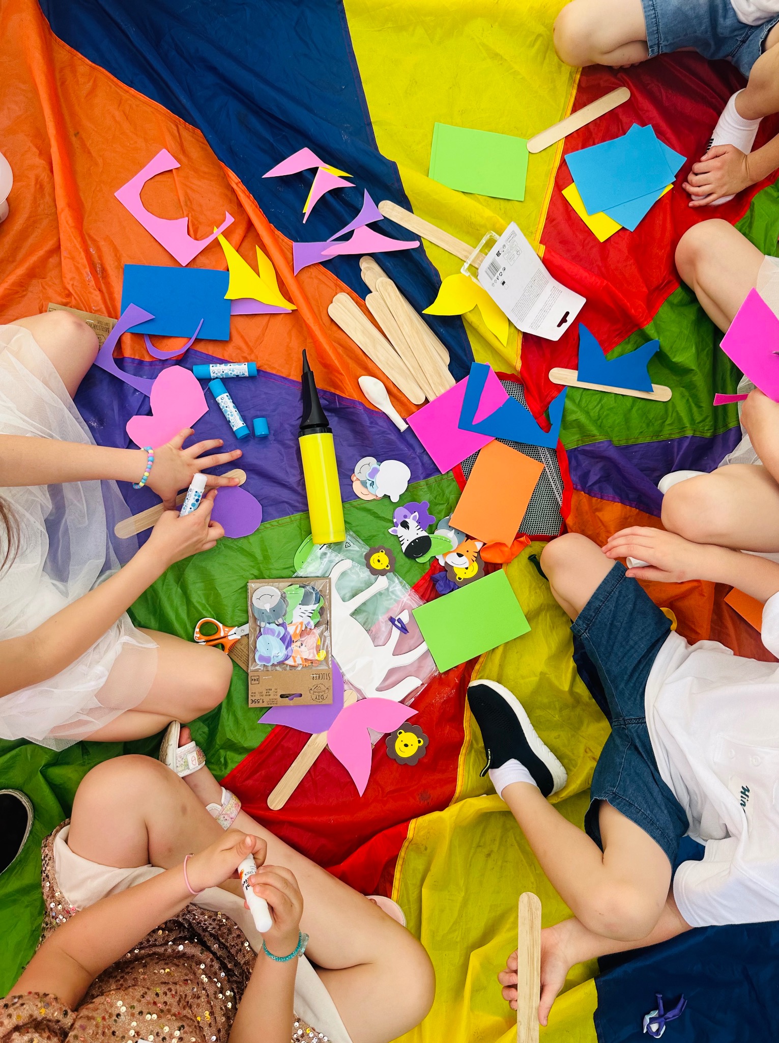 Grupa dzieci siedzi na kolorowej płachcie, tworząc ozdoby z kolorowego papieru, kleju i drewnianych patyczków; widoczne naklejki ze zwierzętami, nożyczki i pompka do balonów.