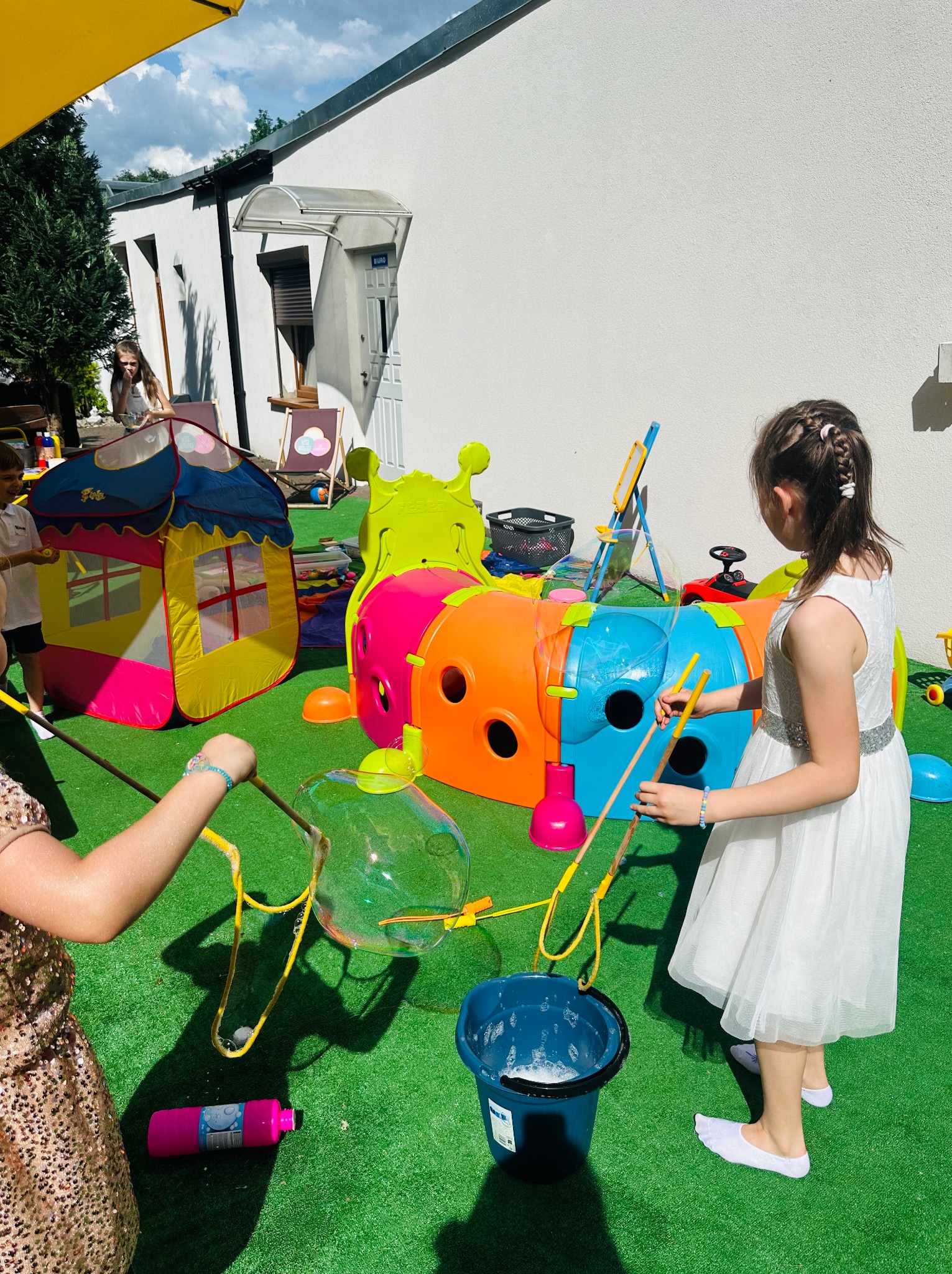 Dzieci puszczają bańki mydlane na trawniku, kolorowy tunel i namiot dla dzieci w tle, widoczne akcesoria do robienia baniek i wiadro z wodą z mydłem.