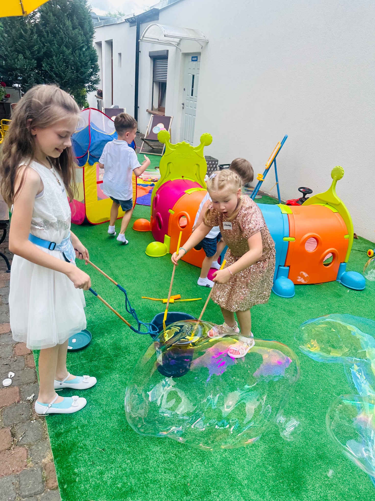 Dziewczynki bawią się w ogrodzie, tworząc ogromne bańki mydlane za pomocą specjalnych kijków i płynu; w tle kolorowy tunel-gąsienica i inne dzieci