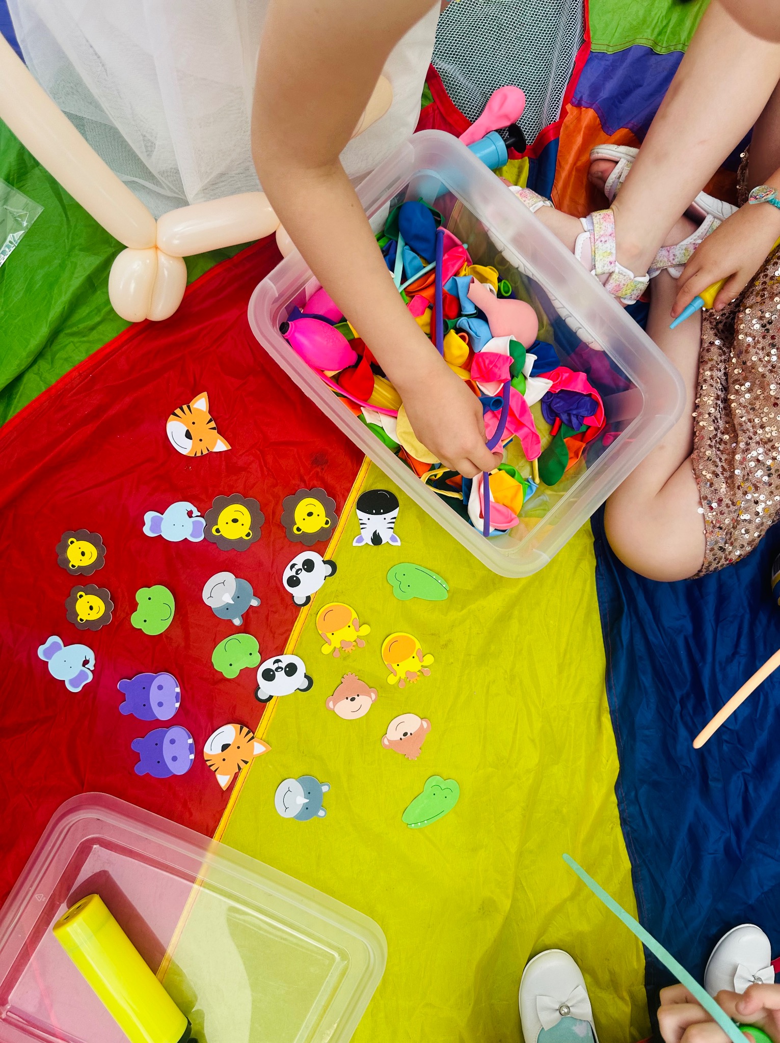 Dzieci siedzą na kolorowej macie, bawiąc się balonami, pompkami i naklejkami ze zwierzętami podczas animacji.