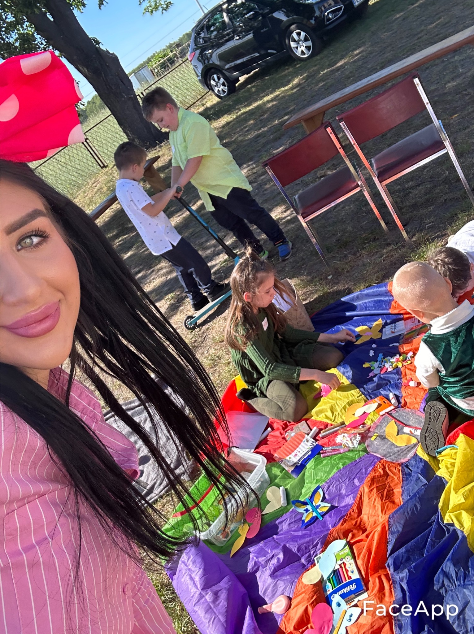 Selfie kobiety z długimi czarnymi włosami w różowej koszuli na tle dzieci bawiących się na kolorowej chuście animacyjnej na trawie, z elementami do animacji i zabaw.