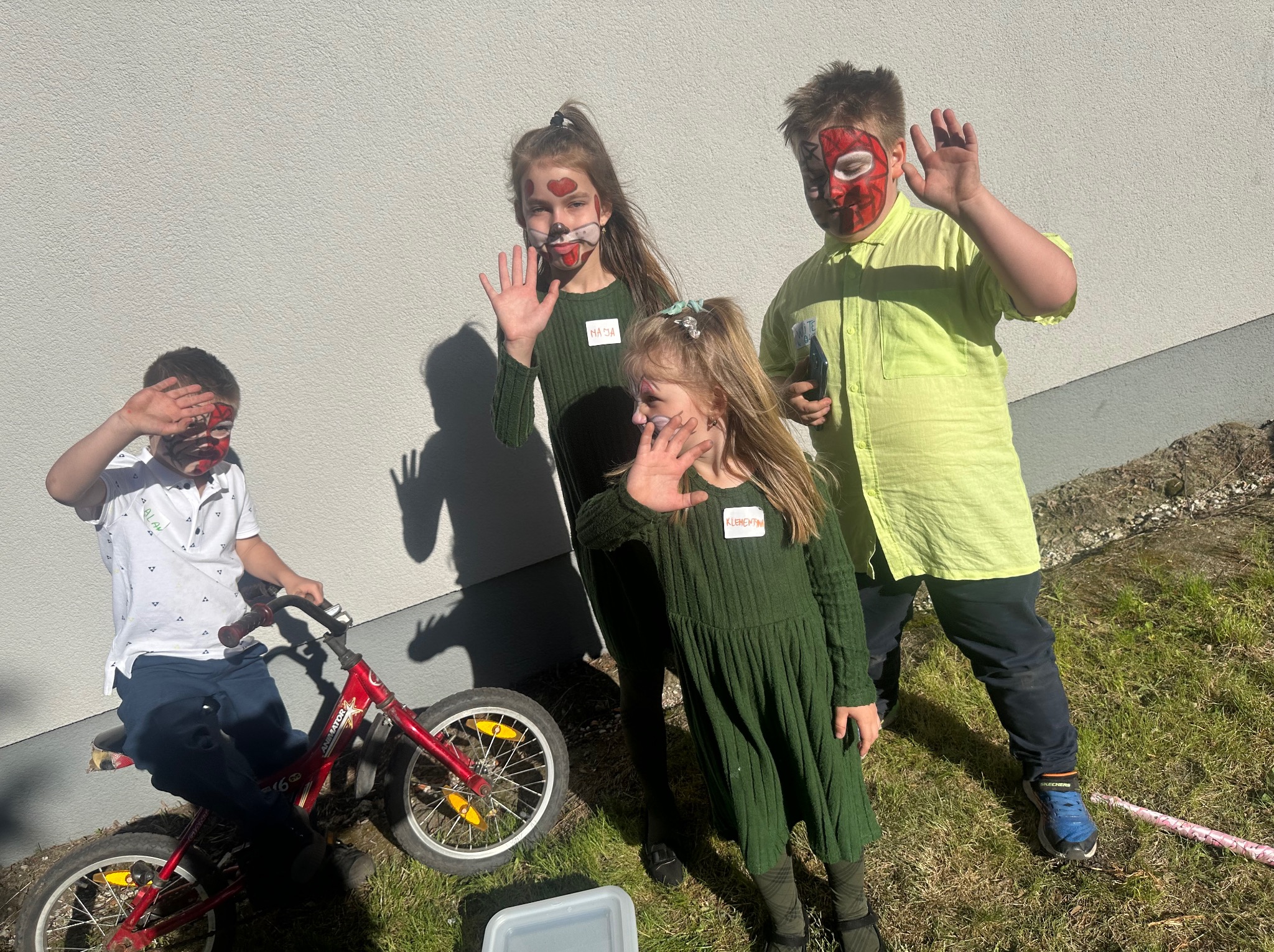 Grupa dzieci z pomalowanymi twarzami, w tym chłopiec na czerwonym rowerze z napisem 'Animator', pozdrawia na tle jasnej ściany i trawy.