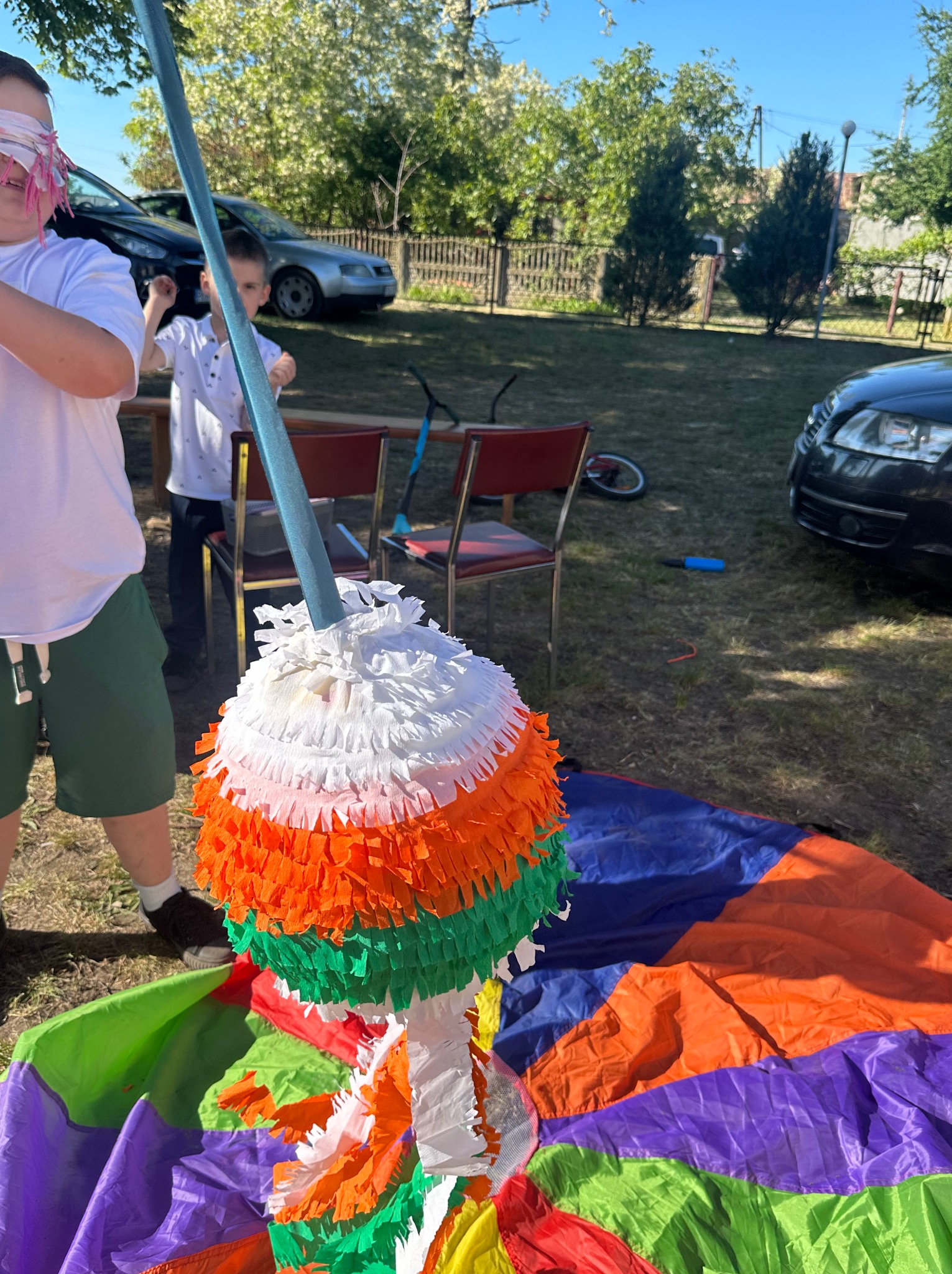 Rozbijanie kolorowej piniaty podczas zabawy na trawie, z dzieckiem w opasce na oczach, w tle samochody i drzewa.