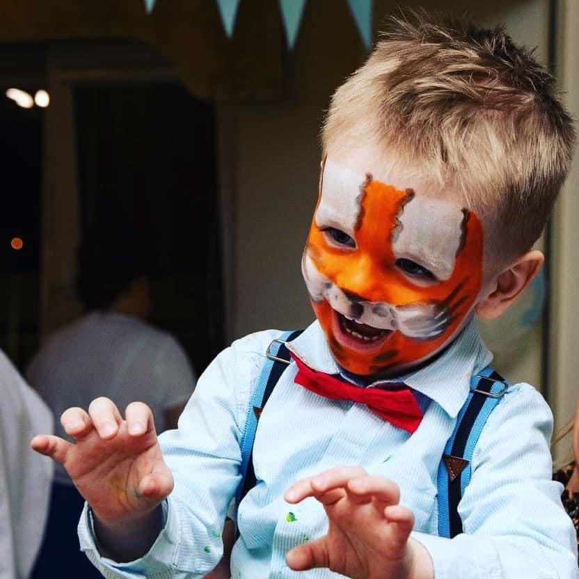 Chłopiec z pomalowaną twarzą w tygrysa, ubrany w koszulę z muchą i szelkami, w Koninie. Radosny wyraz twarzy i uniesione dłonie.