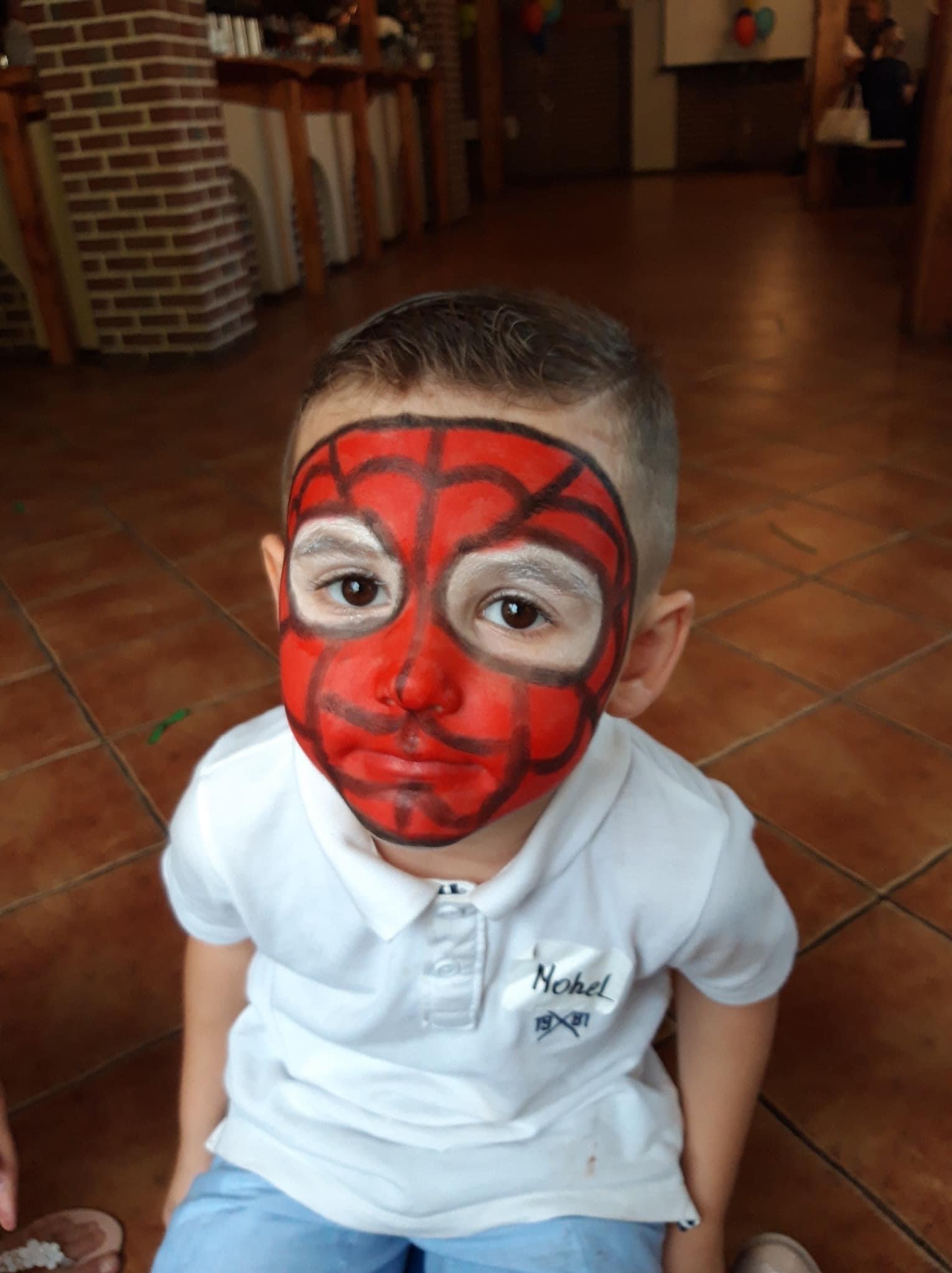 Chłopiec z pomalowaną twarzą na podobieństwo Spider-Mana, w białej koszulce polo z napisem, siedzący na brązowej podłodze z płytek.
