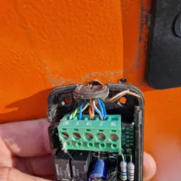 Otwarta obudowa sterownika z widoczną płytką drukowaną, zieloną listwą zaciskową i podłączonymi przewodami na tle pomarańczowej powierzchni bramy.