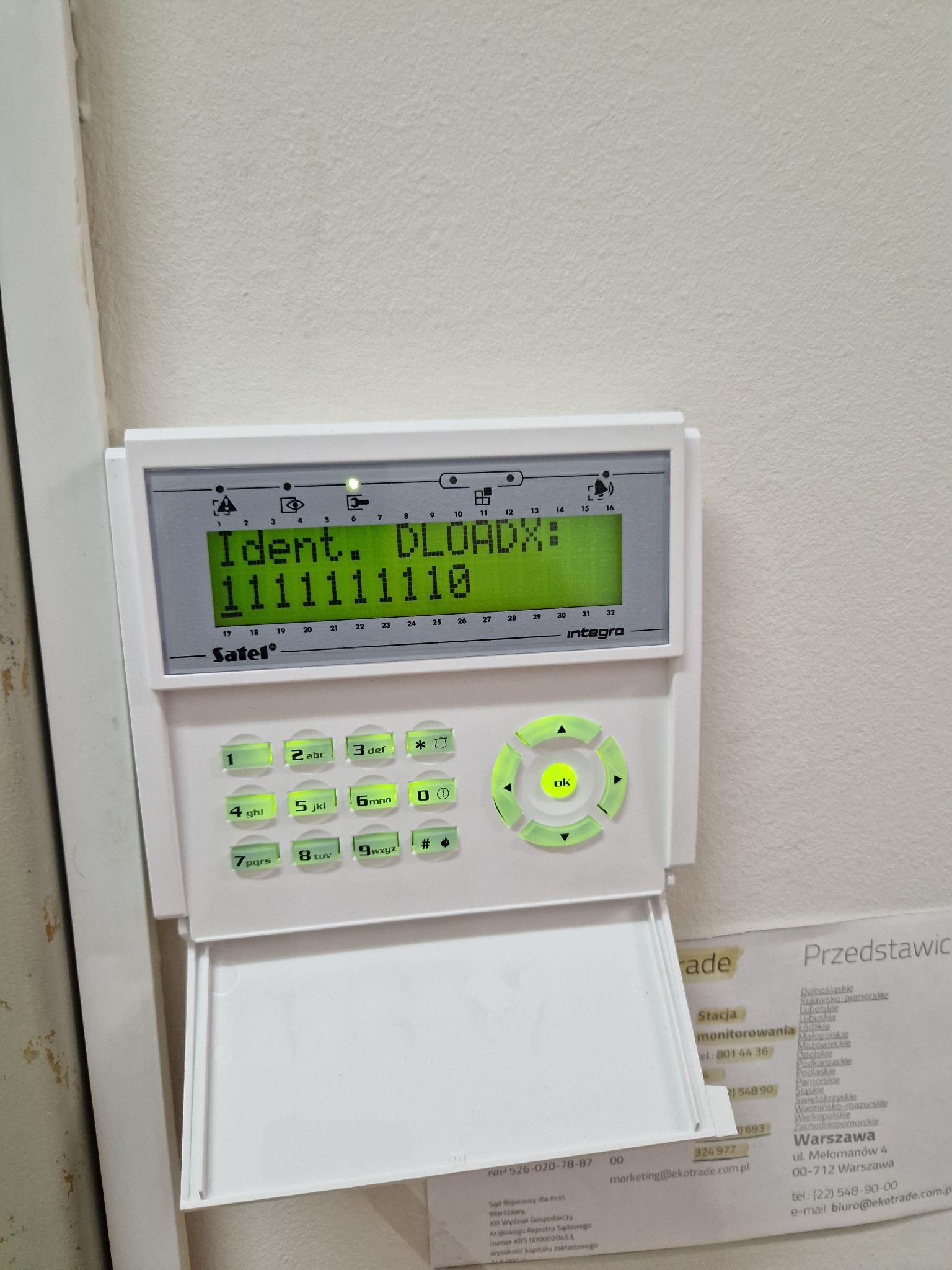 Panel alarmowy Satel Integra z podświetlonym ekranem LCD i klawiaturą numeryczną, zamontowany na jasnej ścianie, widoczny fragment wizytówki firmy z danymi kontaktowymi.