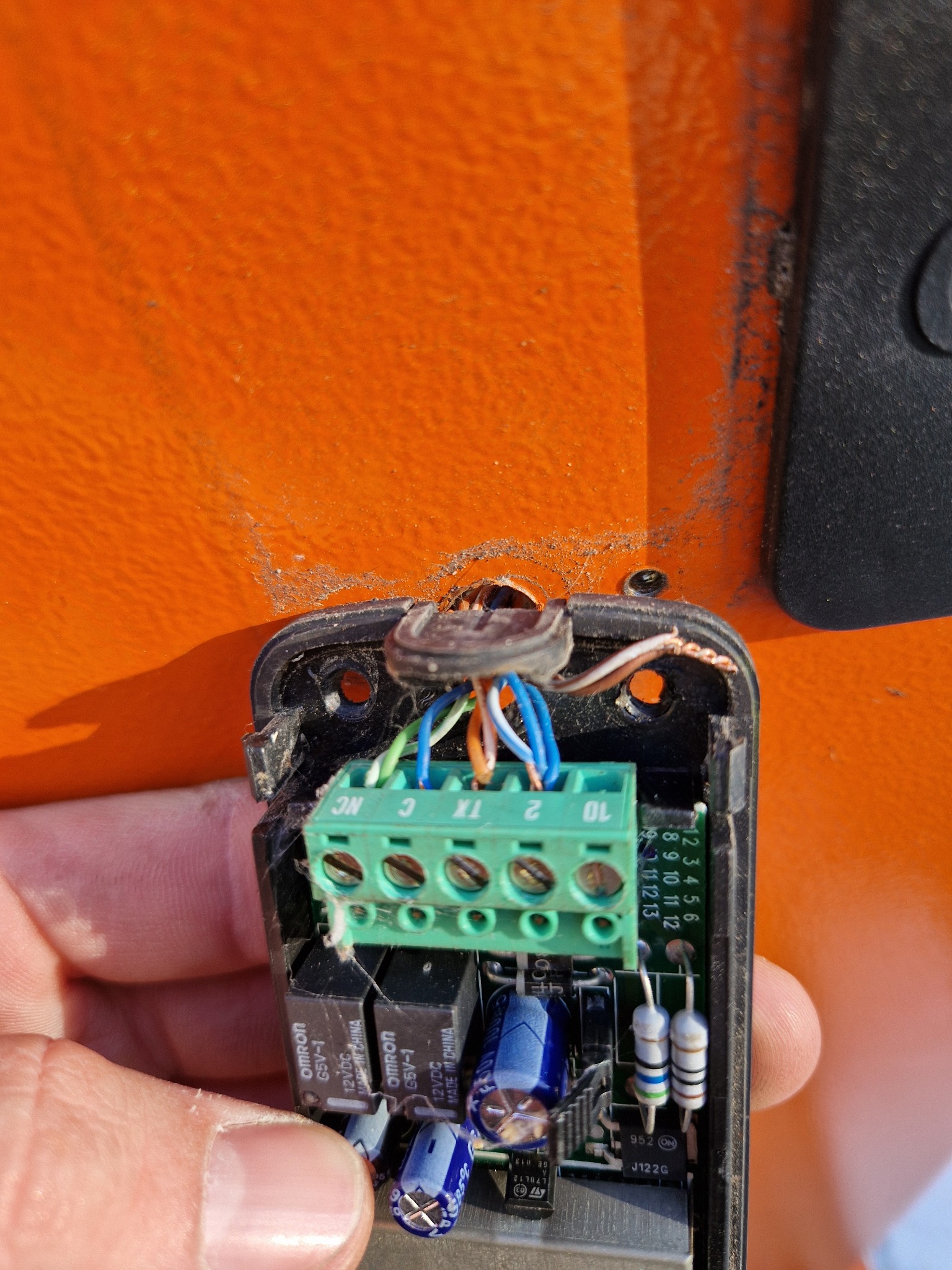 Otwarta obudowa sterownika z widoczną płytką drukowaną, zieloną listwą zaciskową i podłączonymi przewodami na tle pomarańczowej powierzchni bramy.