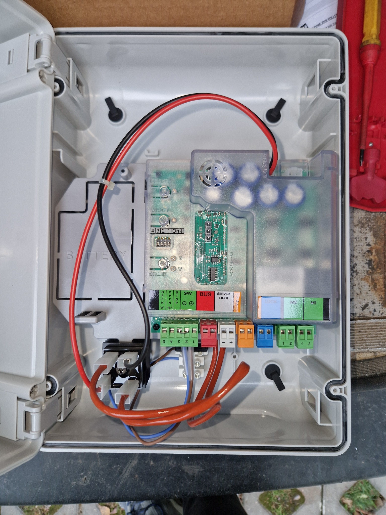 Otwarta, biała skrzynka sterownicza z widoczną elektroniką, przewodami w kolorach czerwonym, czarnym i pomarańczowym, oraz złączkami w kolorach czerwonym, białym, pomarańczowym, niebieskim i zielonym.