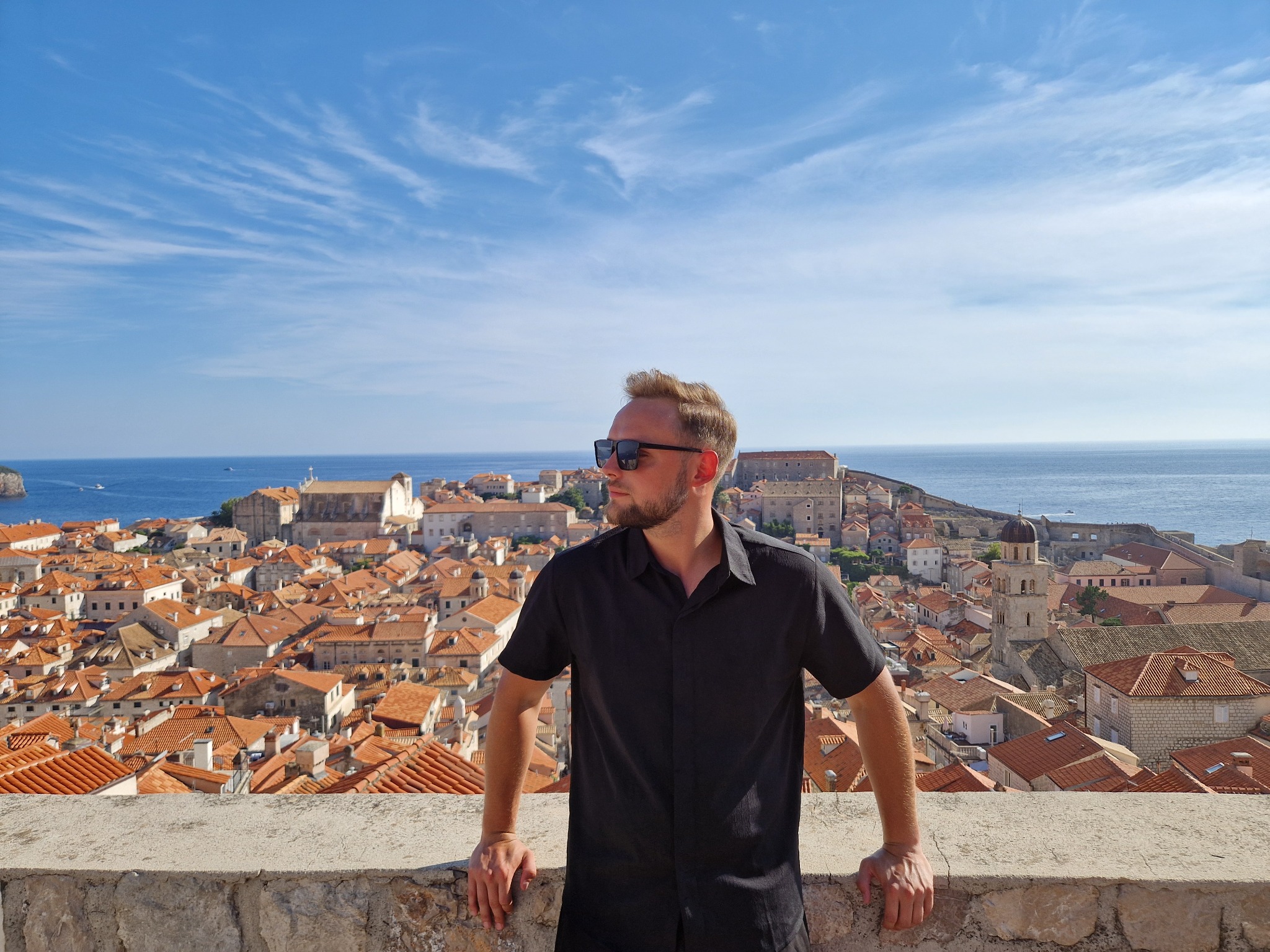Mężczyzna w czarnej koszuli i okularach przeciwsłonecznych opiera się o kamienny mur, z widokiem na miasto z czerwonymi dachami, morze i błękitne niebo z chmurami.